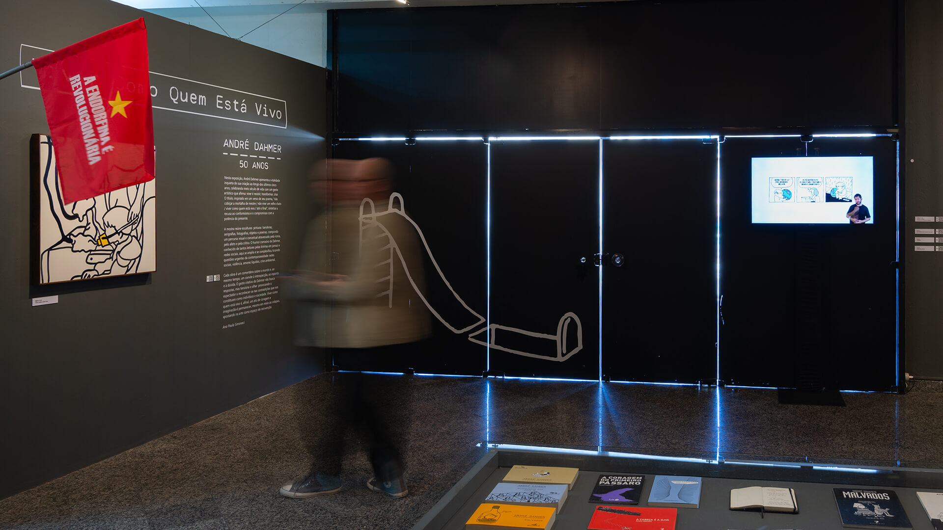 Parede preta com título da exposição e texto informativo, uma pessoa passa em movimento desfocada. Em primeiro plano, na lateral esquerda, uma bandeira vermelha com texto 