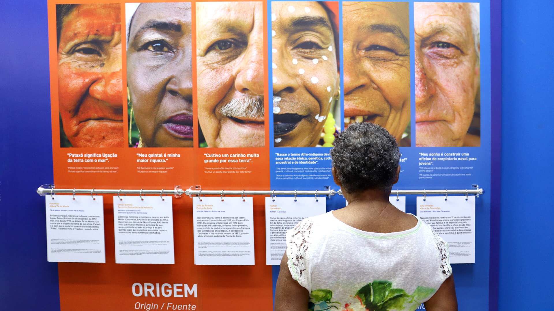 Sob uma parede azul, está um grande painel centralizado na foto, exibindo seis retratos de rostos pessoas diversas, como indígenas, negras e brancas, recortados pela metade. Cada retrato tem um título e um texto abaixo. Na parte inferior a direita, uma mulher de cabelos curto, vestido uma camisa branca e de costas, observa o painel. 