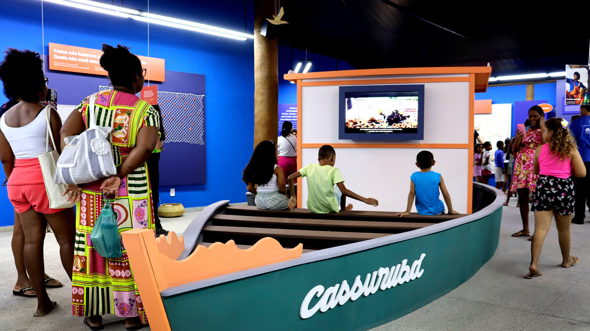 Sala da exposição com paredes azul e um grande barco cenográfico ao centro.  Pessoas circulam pela sala e interagem com o barco e os painéis.