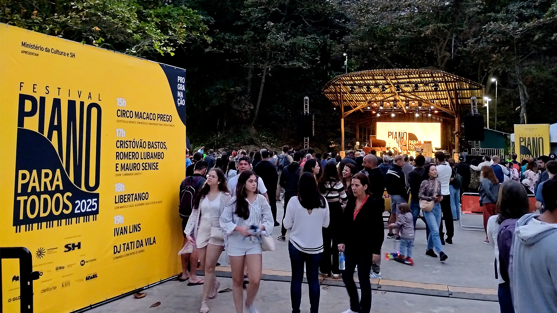 Grande painel amarelo à esquerda com o nome do evento “Festival Piano Para Todos 2025” e a programação impressa em letras pretas. No centro e ao fundo, muitas pessoas caminham e conversam em frente a um palco iluminado. O clima é de fim de tarde, e o público veste roupas leves e casuais.