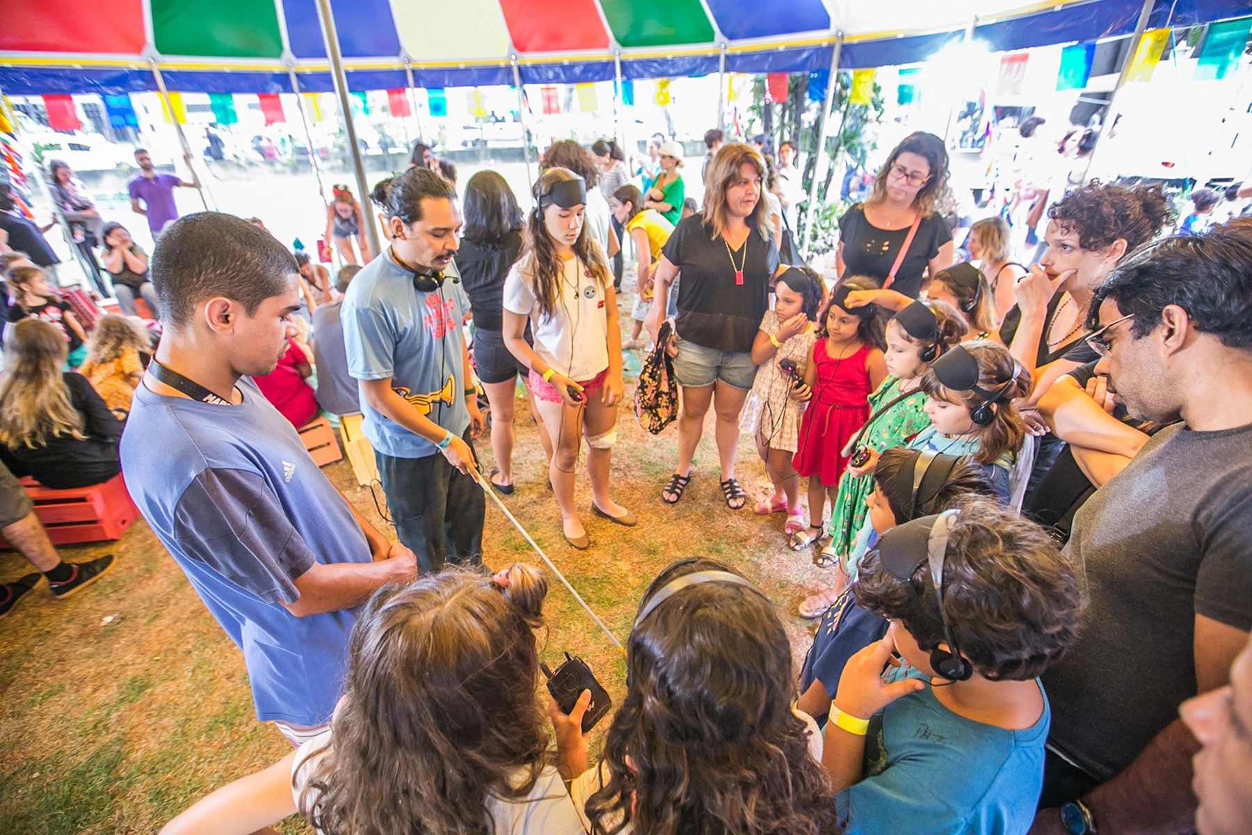 Um grupo de pessoas, incluindo adultos e crianças, reunidas sob uma tenda. Várias crianças estão usando vendas ou fones de ouvido durante uma atividade sobre acessibilidade.