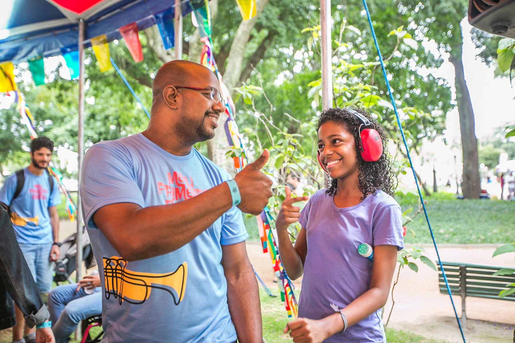 Um homem e uma jovem sorrindo um para o outro e fazendo gestos com o polegar para cima, com a menina usando fones de ouvido de proteção acústica de cor vermelha; Eles estão em ambiente ao ar livre e arborizado, parte de uma tenda azul aparece na foto.