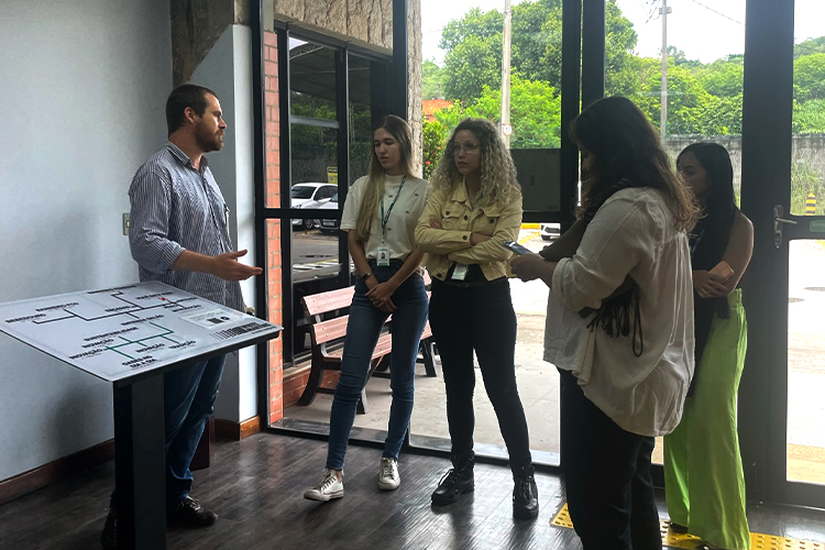 Grupo de pessoas conversando em volta de uma placa de mapa t&aacute;til. Atr&aacute;s delas, uma grande porta de vidro com estrutura de metal preta.