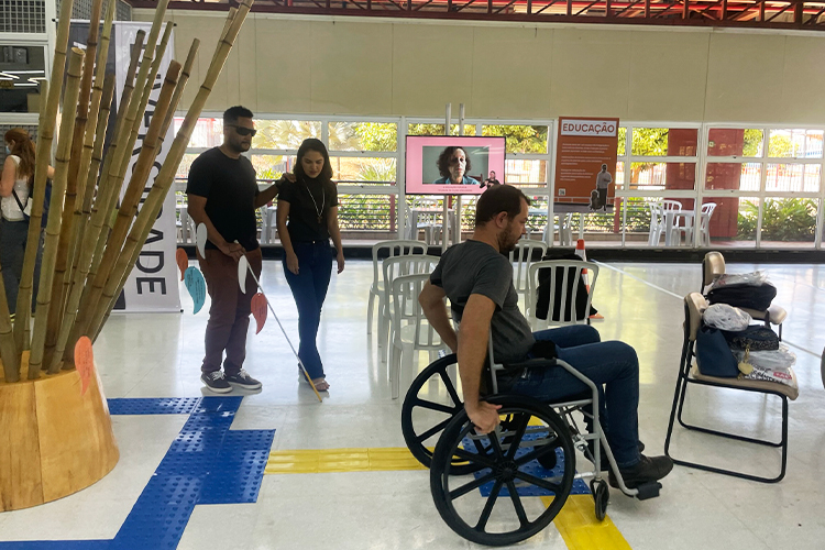 Pessoas fazem simula&ccedil;&otilde;es na exposi&ccedil;&atilde;o, que apresenta recursos de acessibilidade. Em primeiro plano, um homem em cadeira de rodas, atr&aacute;s dele um outro homem usa &oacute;culos escuros e bengala e &eacute; conduzido por uma mulher.