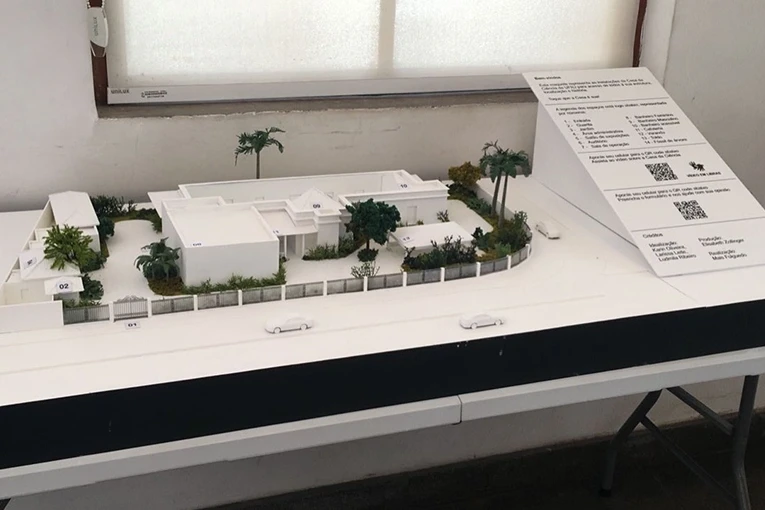 Maquete arquitet&ocirc;nica de um conjunto de edif&iacute;cios com paredes e telhados brancos e vegeta&ccedil;&atilde;o ao redor. Ao lado, um placa de fundo branco, com legenda da maquete, textos e dois QRcodes.