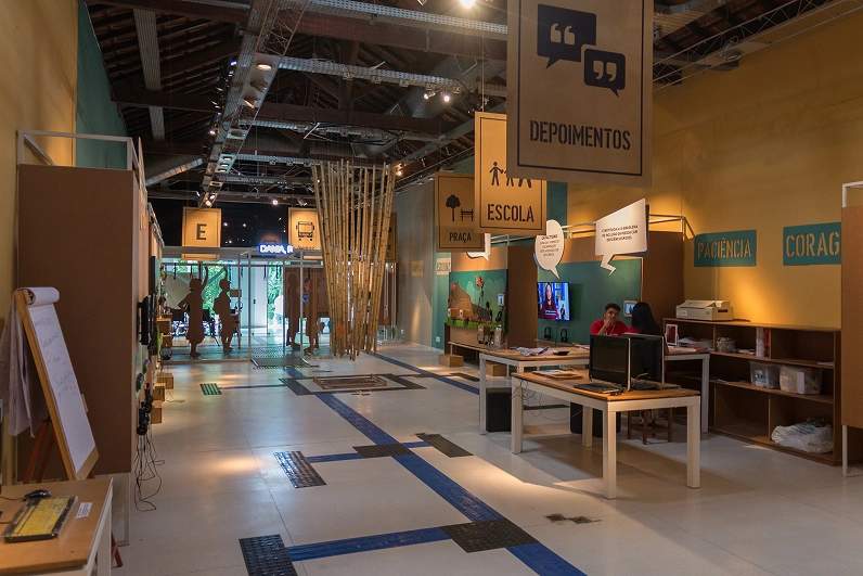 Exposi&ccedil;&atilde;o Cidade Acess&iacute;vel em espa&ccedil;o amplo. Placas de MDF suspensas e penduradas em suportes met&aacute;lico no teto, fazem a sinaliza&ccedil;&atilde;o do local, com nomes e icones. Ao redor do espa&ccedil;o, pain&eacute;is alinhados &agrave;s paredes e no centro um v&atilde;o com percurso delimitado com piso t&aacute;til.