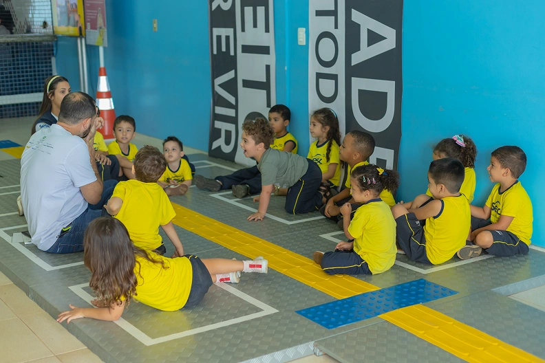 Grupo de crian&ccedil;as vestidas com uniforme, camisa amarela e short azul, sentadas sob &aacute;rea demarcada e com piso t&aacute;til