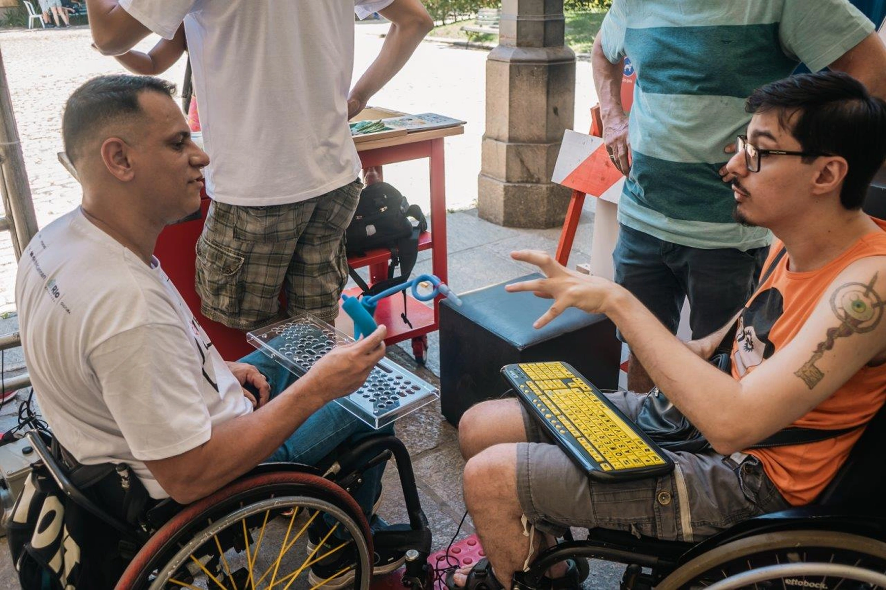 Dois homens, ambos em cadeiras de rodas. Um segura um dispositivo azul e uma placa com furos feita de acrílico, enquanto o outro gesticula com a mão esquerda e tem um teclado adaptado à sua frente.