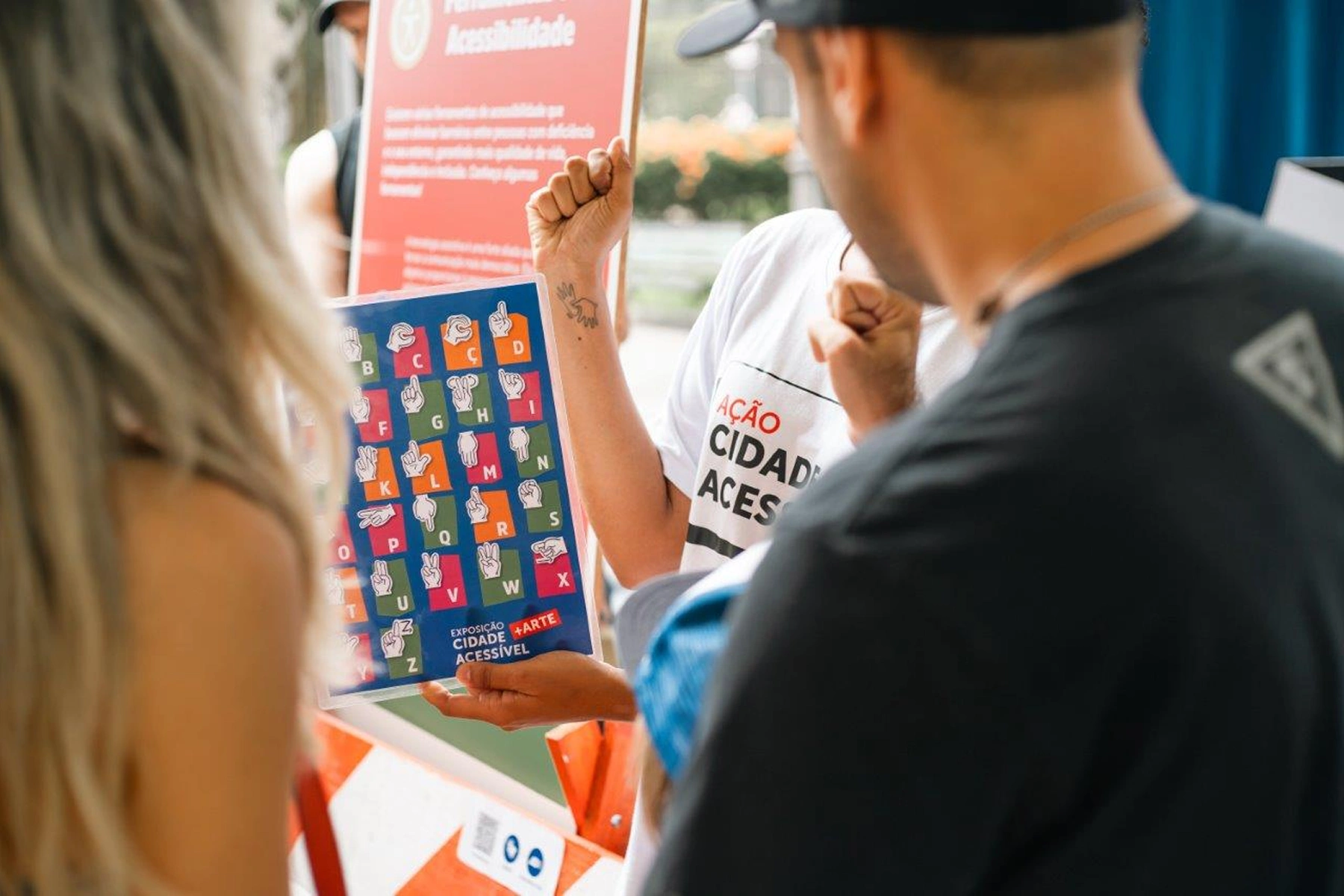 Duas pessoas observam uma tabela de mão do alfabeto em Libras, na foto podemos observar parte de uma mediadora segurando a tabela com a marca 